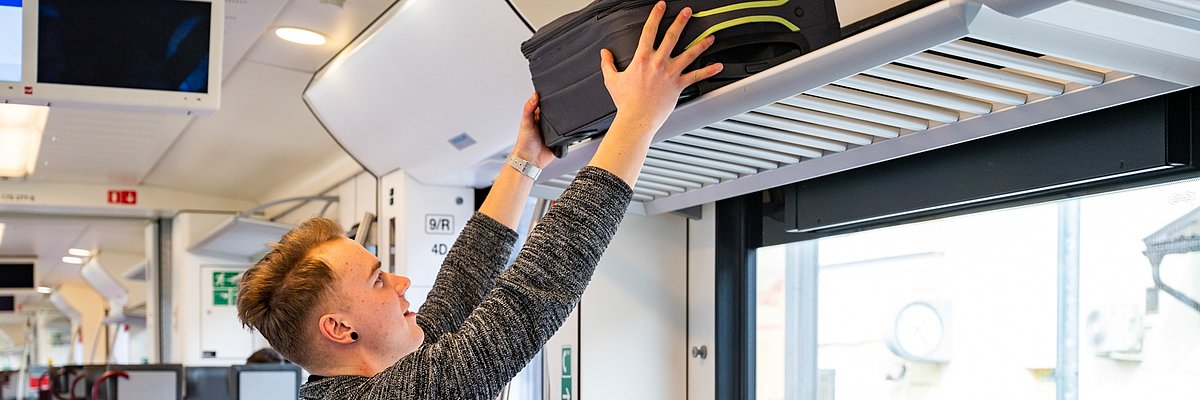 Un giovane passeggero in treno posiziona una valigia sulla cappelliera, mentre una ragazza seduta vicino al finestrino lo guarda.