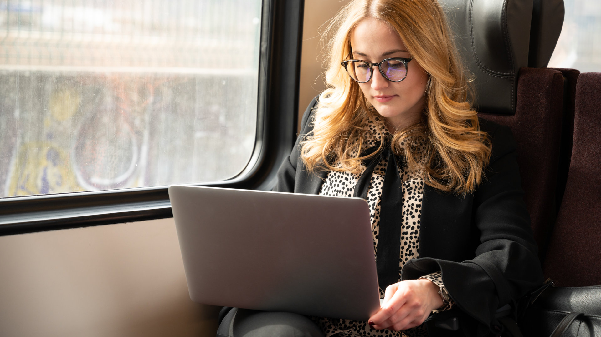 Una ragazza con gli occhiali lavora sul laptop, seduta vicino al finestrino in treno.