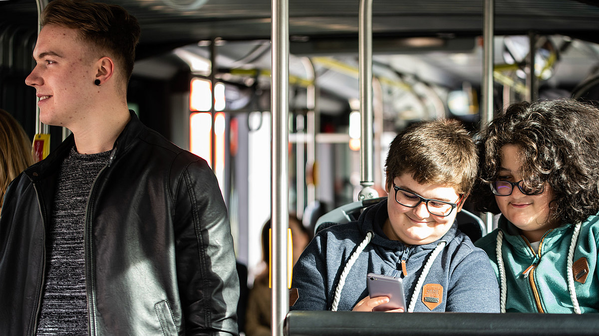 Due ragazzi seduti in autobus controllano insieme uno smartphone. Sulla sinistra un altro ragazzo in piedi che gli volta le spalle.
