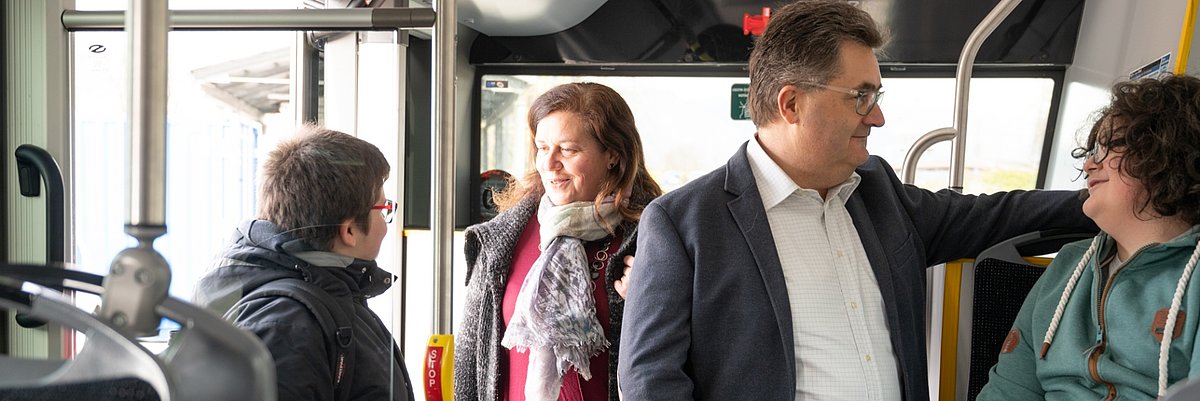 Quattro passeggeri, 2 adulti e 2 ragazzi, conversano su un autobus. Un passeggero è seduto.