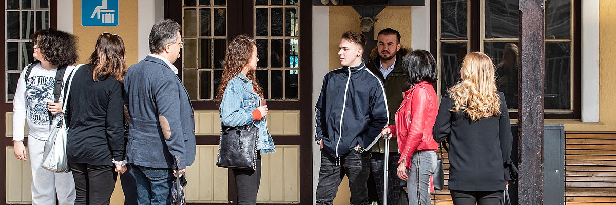 Gruppo di persone in attesa vicino all'edificio di una stazione. Alcuni conversano tra loro.